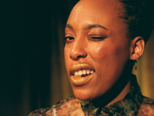 A Close-Up Portrait Of A Black Person Winking At The Camera. They Have Shimmering Gold Lipstick On Their Lips And A Small, Amber-Colored Gummy Held Between Their Teeth. They Are Also Wearing A Nose Ring And A Small Earring Featuring A Hamsa Hand Charm. The Lighting Is Warm And Dramatic, Highlighting Their Skin Against A Dark, Shadowy Background.