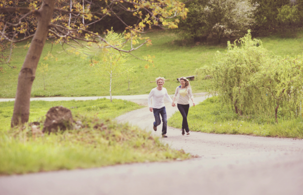 A Young Couple Runs Hand-In-Hand Down A Winding Paved Path In A Lush, Green Park. The Man Wears A Long-Sleeved White Shirt And Blue Jeans, While The Woman Wears A Light-Colored Top And Dark Pants. They Are Both Smiling, Captured In A Joyful Moment With Trees And Grassy Hills In The Background.