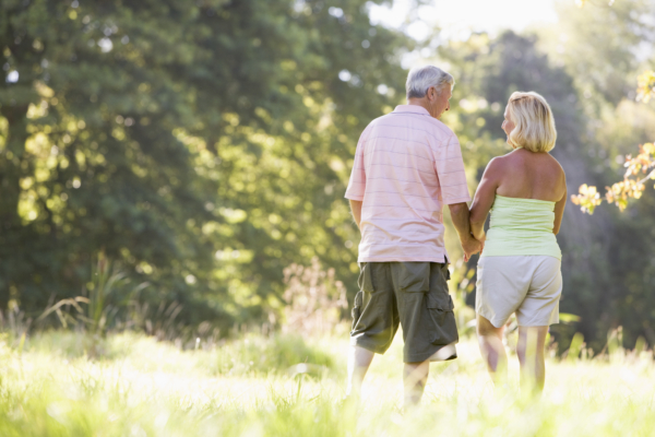 An Older Couple Is Seen From Behind, Walking Hand-In-Hand Through A Sun-Drenched, Grassy Field. The Man Is Wearing A Light Pink Polo Shirt And Olive Cargo Shorts, While The Woman Wears A Lime Green Tank Top And Light-Colored Shorts. They Are Facing Toward A Backdrop Of Lush Green Trees, With Bright Sunlight Filtering Through The Leaves And Creating A Soft Glow Across The Grass.