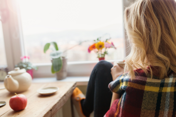 A Blonde Woman Is Seen From Behind, Sitting Comfortably By A Large Window And Looking Out At A Bright, Blurred Cityscape. She Is Wrapped In A Red, Yellow, And Blue Plaid Blanket And Holds A Mug In Her Hands. In The Foreground, A Wooden Table Holds A White Teapot, A Small Saucer, And A Red Apple. On The Windowsill, A Potted Orchid And A Small Vase Of Colorful Gerbera Daisies Add A Touch Of Nature To The Cozy Indoor Setting.